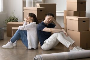 Tired frustrated married couple sitting back-to-back on floor surrounded by cardboard moving boxes